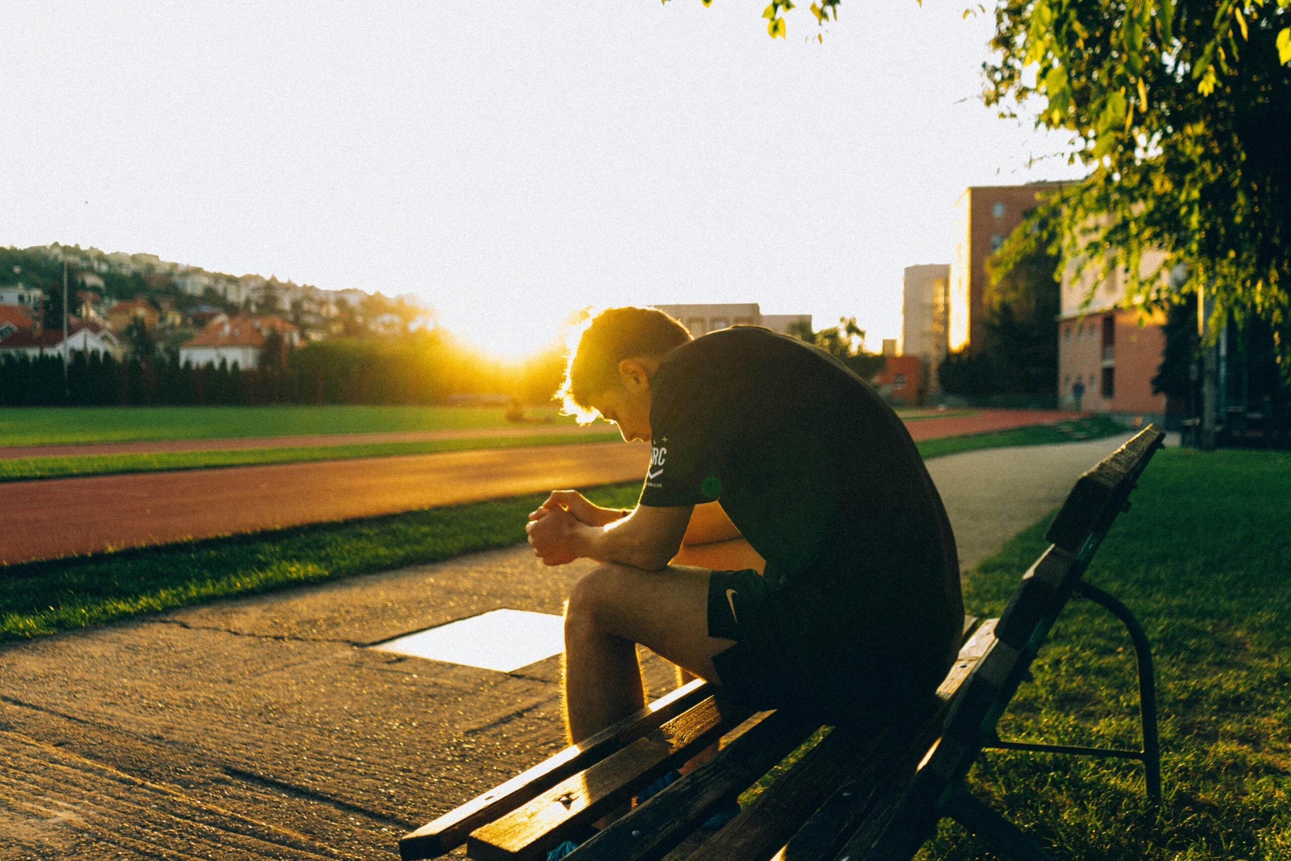 Person sitting on bench during sunset, athletic field nearby.